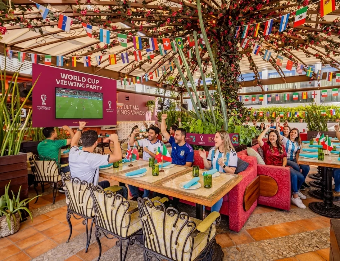 A group of people excitedly watch a World Cup soccer match on a large screen at an outdoor restaurant decorated with international flags. They wave flags, cheer, and sit around tables with food and drinks.