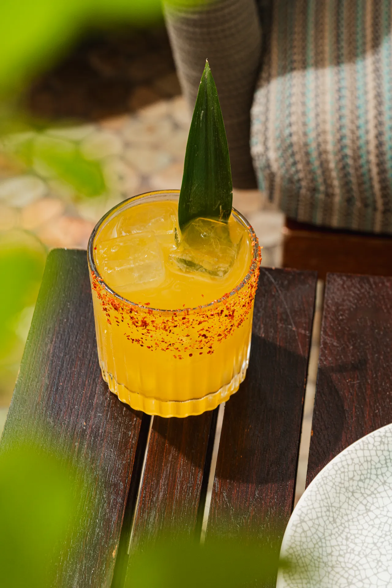 A glass filled with orange cocktail on ice, rimmed with chili powder, and garnished with a pineapple leaf, sits on a wooden table with a patterned chair and a white plate nearby.